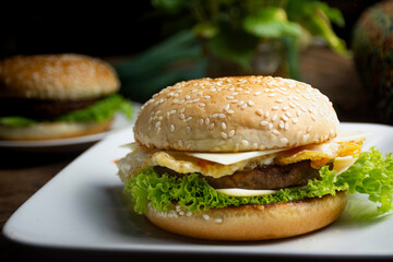 burger with lettuce, cheese, tomato, egg, and beef placed on a white plate with a wooden bottom