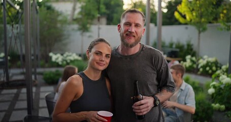 Portrait of a happy blond guy with a beard in a gray T-shirt who hugs his girlfriend and poses while relaxing with company in the courtyard of a country house