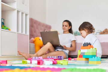 cute little baby girl playing with toys blocks and mother sitting on floor with laptop in room