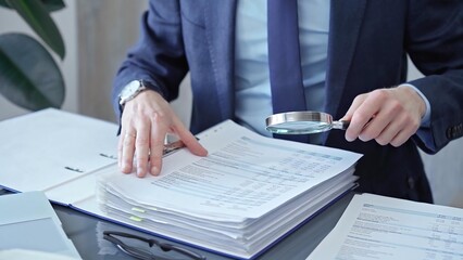 Adult businessman or auditor examining documents with magnifying glass. Professional in a suit scrutinizes financial reports, detail-focused work concept