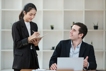 A woman and a man are sitting at a desk with a laptop and a notebook