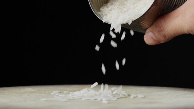 Hand Pouring Uncooked White Rice (Oryza sativa) On A White Surface. Slow Motion (Close-Up) Isolated On Solid Black.