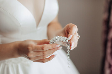 A woman is holding a necklace with pearls and diamonds. The necklace is white and has a gold chain. The woman is wearing a white dress and is standing in front of a curtain