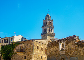Obraz premium The Basilica de la Encina tower in Ponferrada, Spain, rises above historic stone buildings under a clear blue sky, with people walking nearby, showcasing a blend of