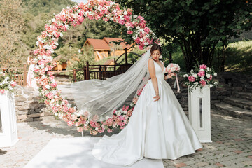 Fototapeta premium A bride is standing in front of a large archway with pink flowers. She is wearing a white dress and holding a bouquet. The scene is set in a garden, with a house in the background