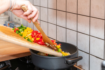 A cooking pot on gas fire. Person putting fresh cut paprikas from a wooden cutting board into a...