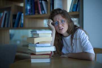 beautiful sad woman student in glasses, sitting with books in library. concept education, university, college, admission, exams