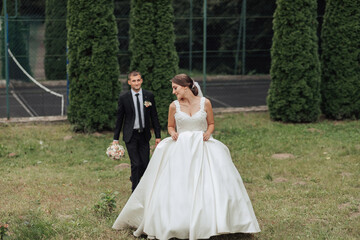Fototapeta premium A bride and groom are walking together in a park. The bride is wearing a white dress and the groom is wearing a suit. They are holding hands and smiling, indicating that they are happy
