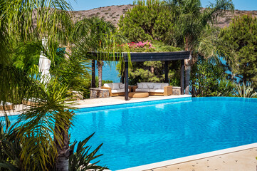 Luxurious swimming pool area surrounded by lush greenery, creating tranquil and serene environment. Pool complemented by covered lounge space featuring cozy sofas and plush cushions, perfect for relax