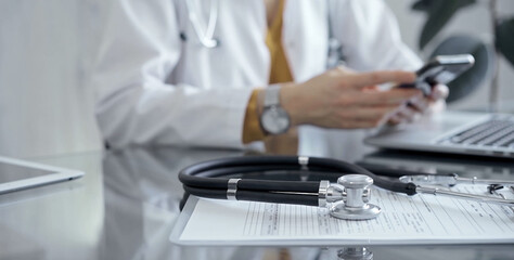 Close-up of doctor's desk with stethoscope and clipboard. Physician is using a tablet at the background. Medicine and health care