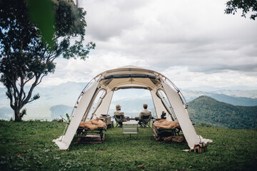 A couple sitting in a tent in the woods with a view of mountains and trees on green grass and blue...