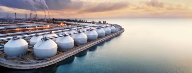 A wide shot of an industrial port with large storage tanks by the water at dusk, with smoke rising from the background.