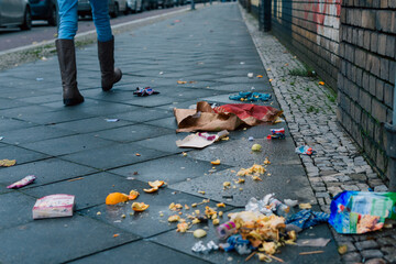 Leftover food and garbage on a sidewalk. A person moves out of the picture, boots