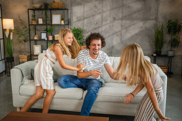 Obraz premium Daughters tickling their father on sofa at home have fun time together
