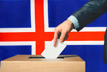 A hand holds a ballot in a ballot box against the background of the Iceland flag. People cast...