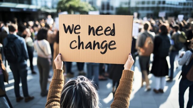 A person holding a sign with the text "we need change" at a peaceful protest, a crowd of people in the background in a city square.