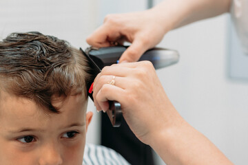 A female hairdresser cuts a little boy's hair with a hair trimmer. Close-up.