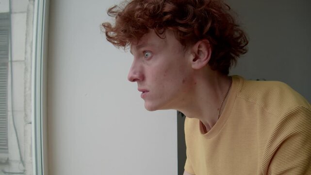 Side chest up shot of young rageful Caucasian man in mustard t-shirt looking furiously out of window and clenching fists, feeling distraught and revengeful indoors