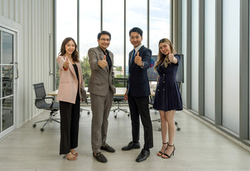 Four people stand together in a bright office with big windows on a sunny day, lifted a finger thumb up with confidence