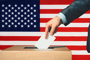 A hand holds a ballot in a ballot box against the background of the US flag. People cast ballots...