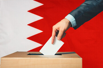 A hand holds a ballot in a ballot box against the background of the Bahrain flag. People cast...