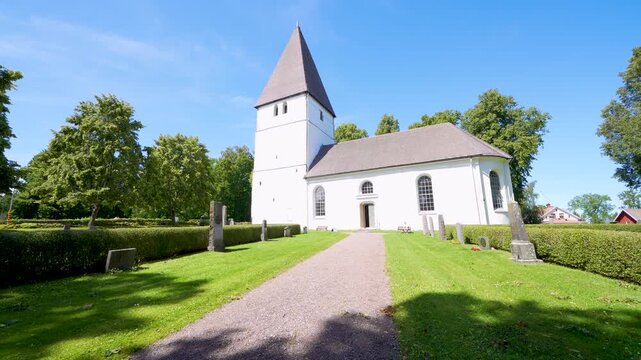 A serene white church with a tall steeple stands on the idyllic grounds of Bjalbo, Sweden, against a clear blue sky.