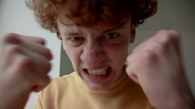 Closeup portrait of rageful young Caucasian man with red curly hair clenching fists and looking at camera, ready to punch, feeling angry and furious indoors