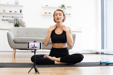 Fototapeta premium Woman in black sportswear practicing yoga at home. Sitting on yoga mat in living room, using phone for online class. Home workout, mindfulness, relaxation, digital fitness.