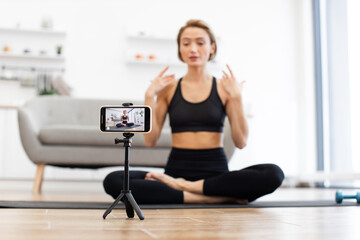 Yoga instructor sitting cross-legged on mat recording virtual fitness class using smartphone. Instructor demonstrating poses in modern living room. Concept of online fitness and digital wellness.
