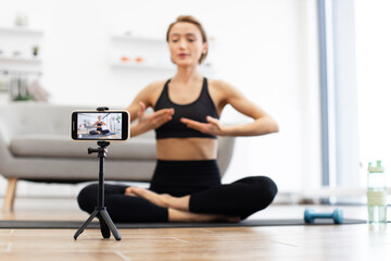 Naklejka premium Woman practicing yoga at home while filming online class on smartphone camera. Wearing black sportswear, sitting cross-legged on mat.