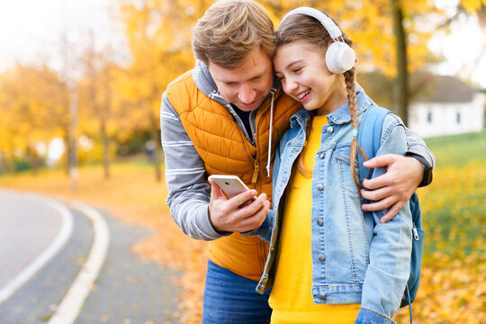 Father and daughter using smartphone on autumn street. Happy kid and parent have fun with gadget outdoors together. Teenage girl in headphones listening to music, laughing, smiling. Lifestyle moments