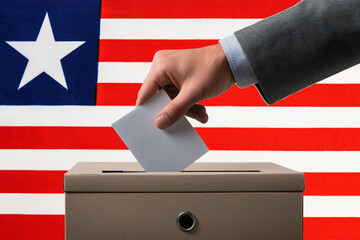 A hand holds a ballot in a ballot box against the background of the Liberia flag. People cast...