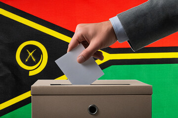 A hand holds a ballot paper in a ballot box against the background of the Vanuatu flag. People cast...