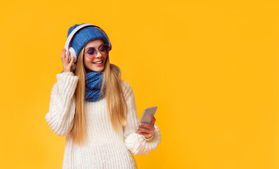 Lifeful girl in sunglasses and blue winter hat watching videos online, using phone and wireless headset, panorama with copy space