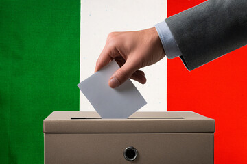 A hand holds a ballot in a ballot box against the background of the Italian flag. People cast...