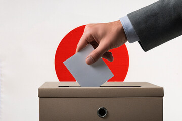 A hand holds a ballot in a ballot box against the background of the Japanese flag. People cast...