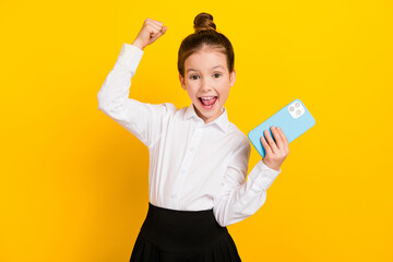 Photo of cute schoolgirl hold phone raise fist wear uniform isolated on yellow color background