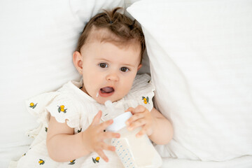 a baby with a bottle of milk or formula in his hands in a bright bedroom, a space for text, a baby food concept, a small child sucking a pacifier with a bottle