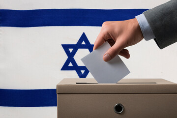 A hand holds a ballot in a ballot box against the background of the Israeli flag. People cast...