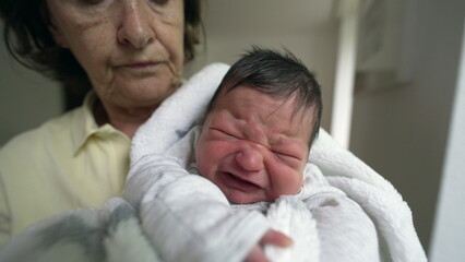 Obraz premium Concerned grandmother holding a fussy newborn baby wrapped in a white blanket. scene captures the love and care between them in a cozy home environment