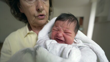 Concerned grandmother holding a fussy newborn baby wrapped in a white blanket. scene captures the love and care between them in a cozy home environment