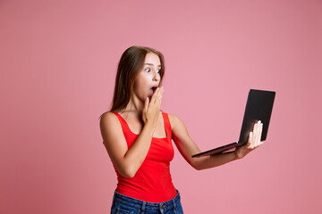 Naklejka premium Portrait of young girl holding laptop with shocked emotional face against pink studio background. Online communication, news, social media. Concept of human emotions, modern lifestyle