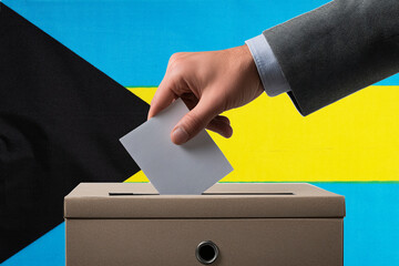A hand holds a ballot in a ballot box against the background of the Bahamas flag. People cast...