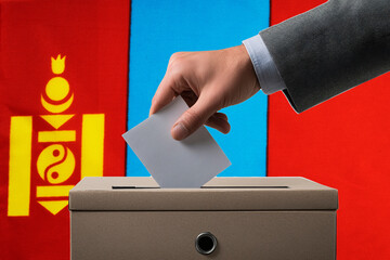 A hand holds a ballot in a ballot box against the background of the Mongolia flag. People cast...