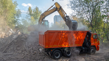 An excavator loads garbage into the back of a large dump truck. Demolition of an old wooden house. A dump truck loads the remains of a destroyed old house into a dump truck. Road equipment.