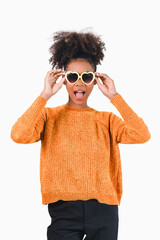 Stylish african american woman in orange sweater wearing heart-shaped sunglasses, making a surprised expression. Isolated on white background.