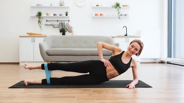Woman performing leg exercises on yoga mat at home using resistance band. She is smiling and appears to be focused on her fitness routine. Home setting includes modern furniture and decor.