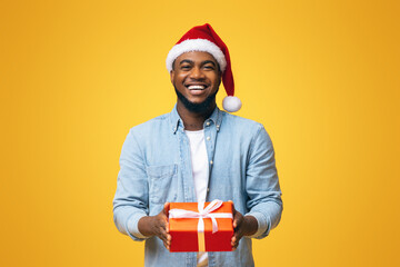 Black Santa. Smiling african american man in red Christmas hat holding xmas gift box over yellow studio background with free space