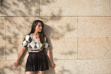 A young, natural woman with brown hair, in a crocheted sweater and black skirt, with depression and anxiety, on a wall with shadows of branches in the background. Concept naturalness, depression.