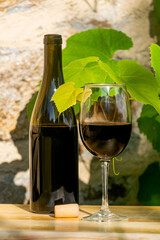 A bottle and a glass of red wine on a wooden table. Stone wall and the leaves of a vine in the background. Copyspace